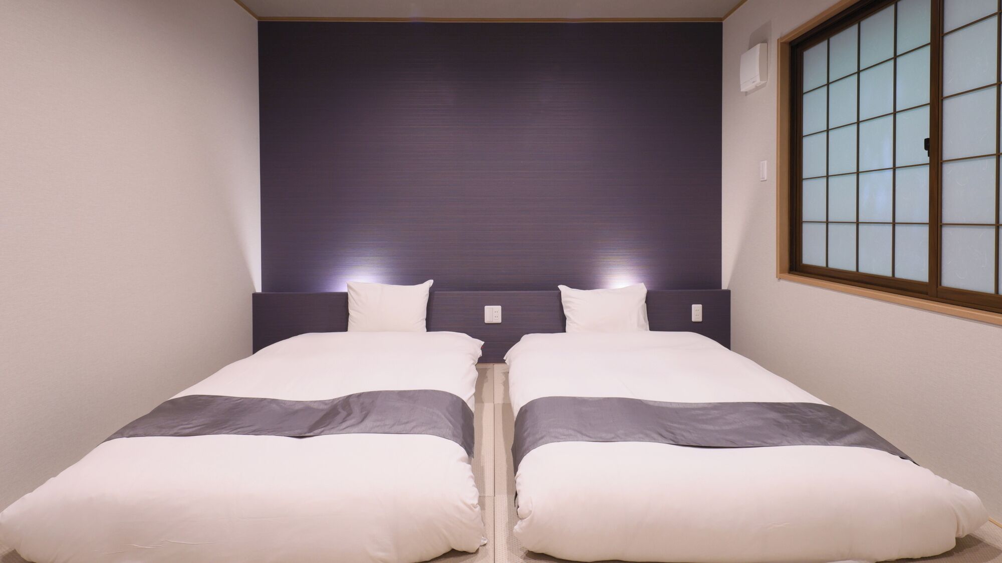 A twin room with two beds, a dark headboard, a window with a grid pattern, and a wall-mounted air conditioner.