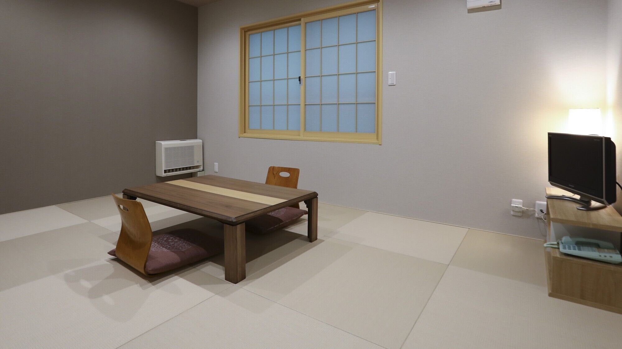 A room with a wooden table, two chairs, a window with blinds, a wall-mounted air conditioner, a TV on a stand, and a lamp.