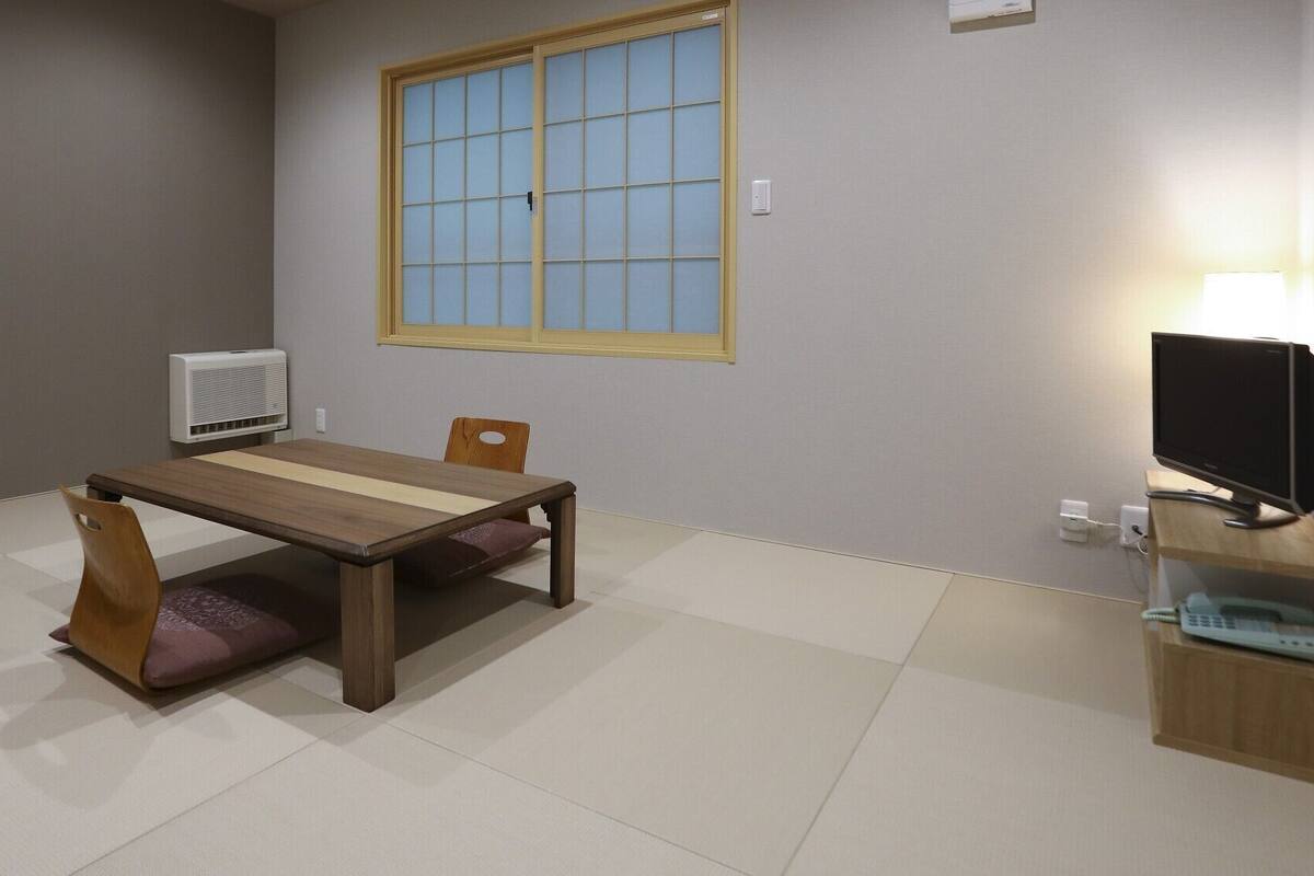 A room with a wooden table, two chairs, a window with blinds, a wall-mounted air conditioner, a TV on a stand, and a lamp.