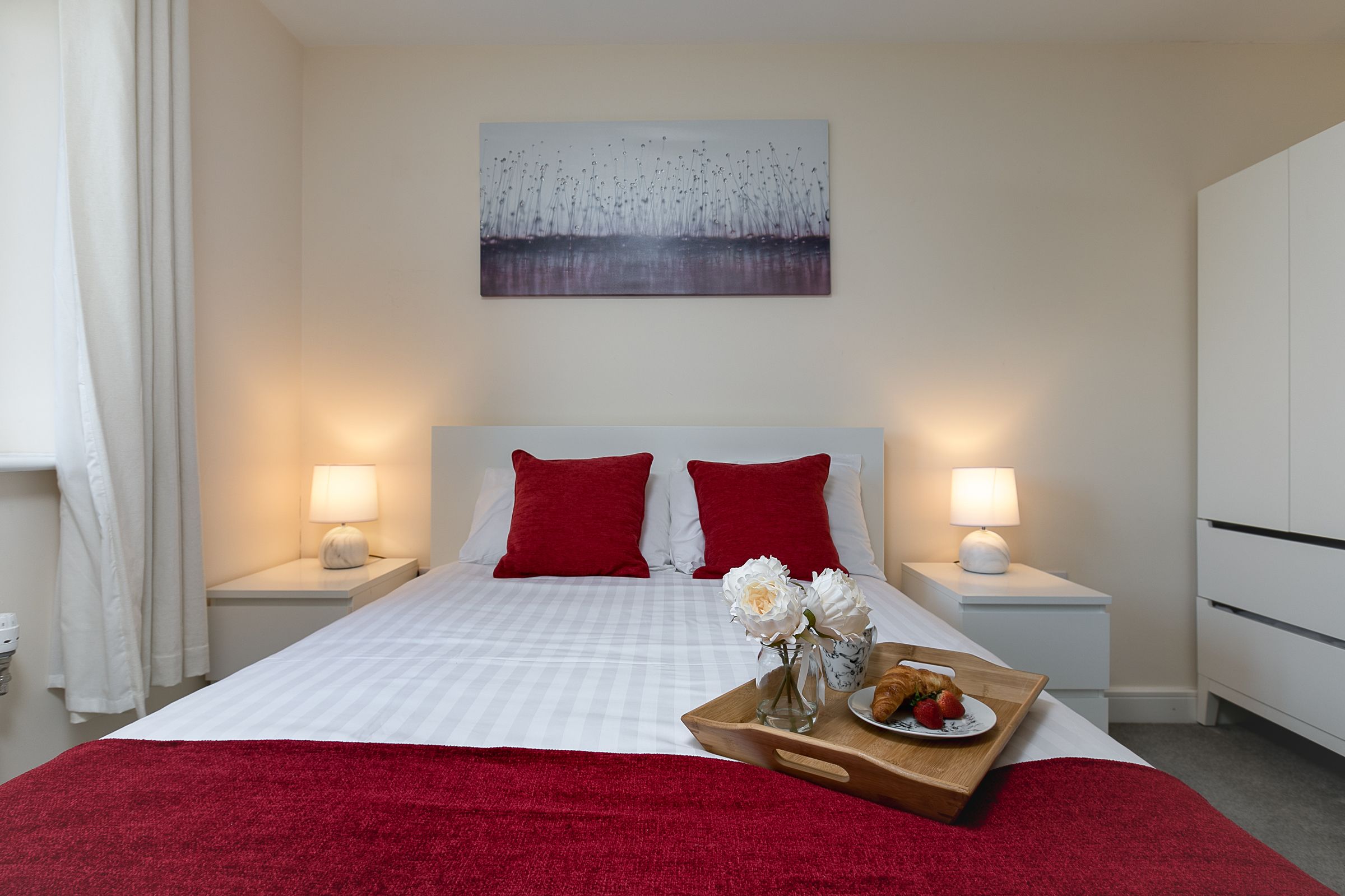 Une chambre d’hôtel avec un grand lit, des oreillers rouges, deux lampes de chevet, un tableau au mur et un plateau avec des fleurs et un croissant posé sur le lit.
