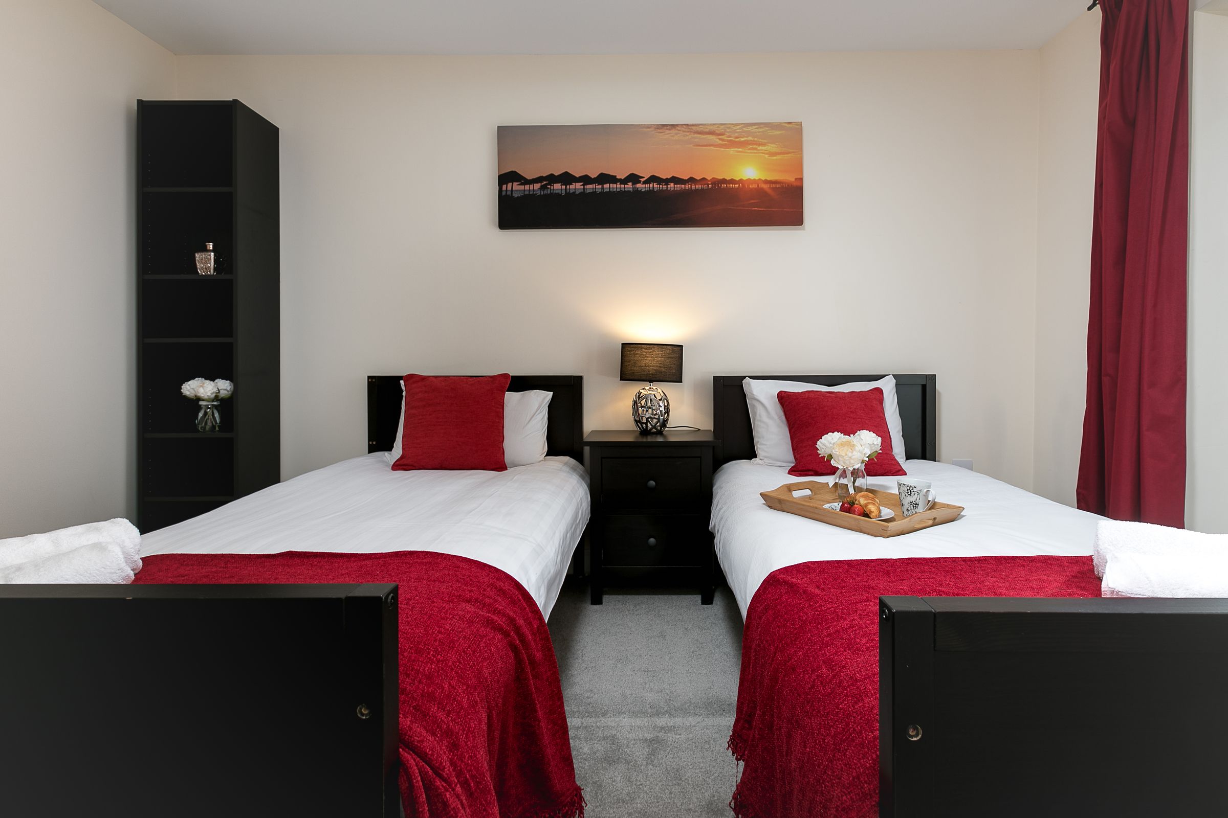 Ein Hotelzimmer mit zwei Betten, einem Nachttisch dazwischen, einem Bild an der Wand und einem roten Vorhang.