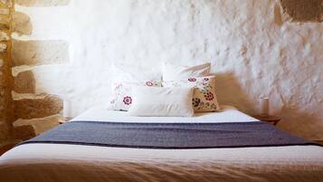 Una cama con ropa blanca y almohadas estampadas, una manta de color oscuro y una pared blanca con ladrillo visto.