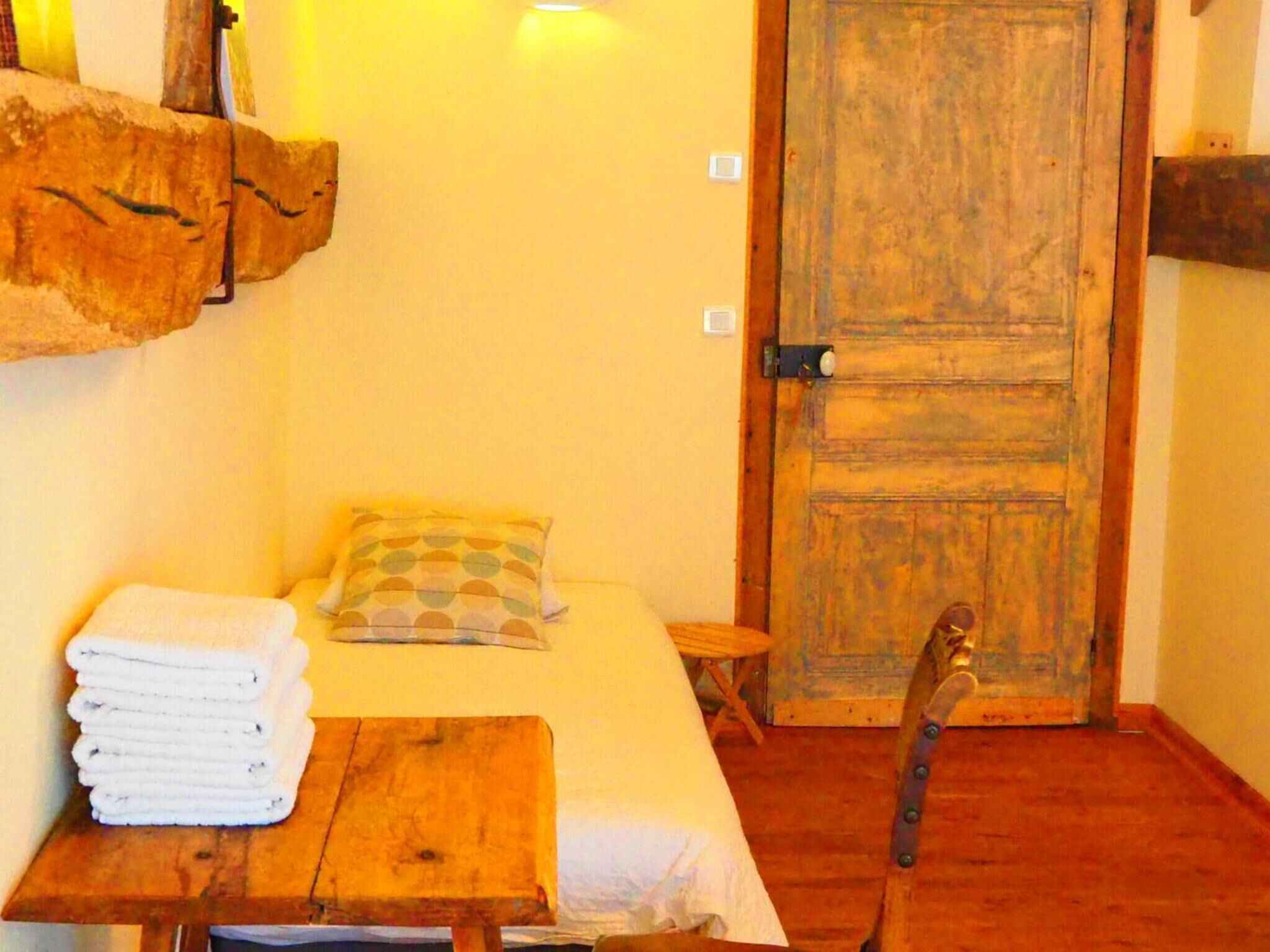 Une petite pièce chaleureuse, avec un lit, une table en bois sur laquelle sont posées des serviettes, une chaise et une porte en bois rustique.
