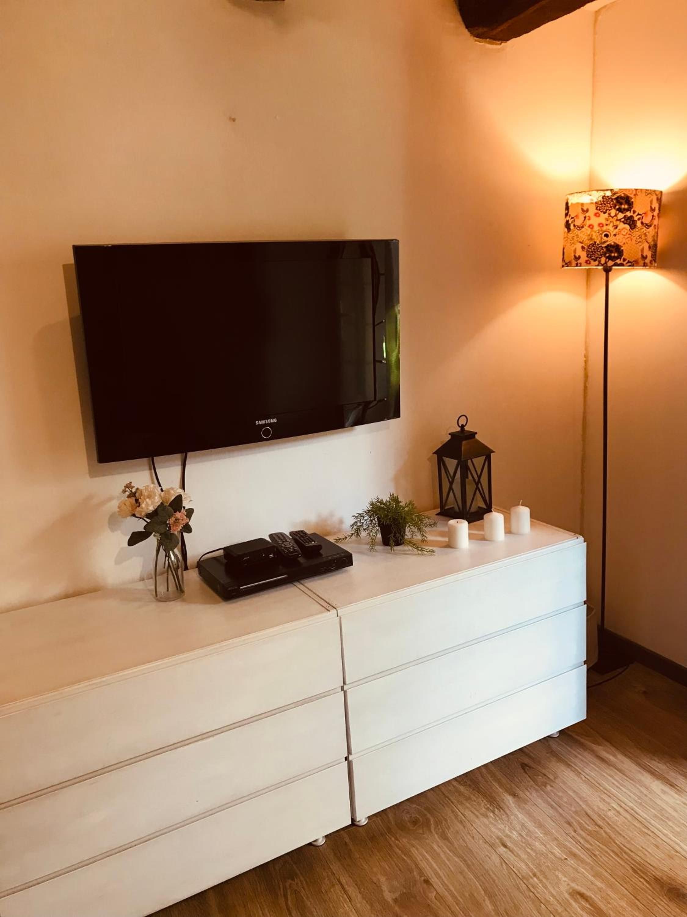 A flat-screen TV mounted on a white cabinet with a remote control, a decorative vase, and a small plant.