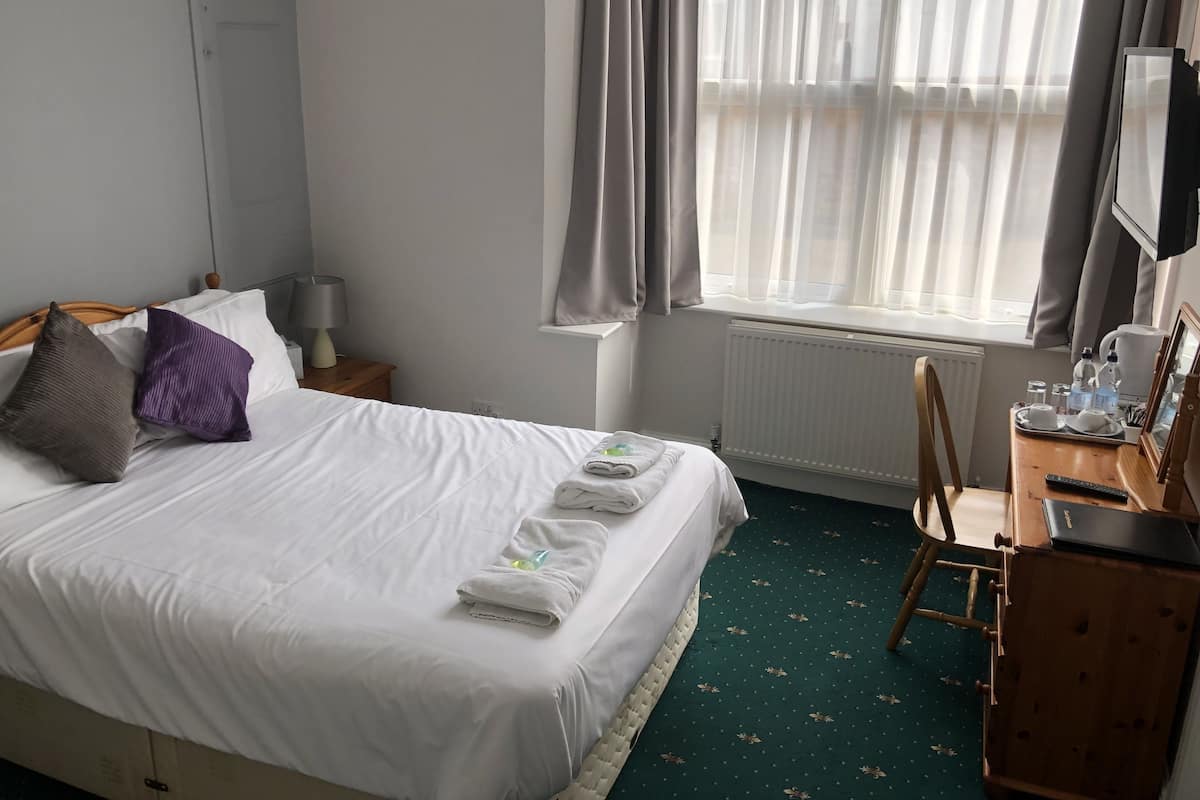 A hotel room with a bed, a desk with a chair, a window with curtains, a radiator, and a television on the wall.