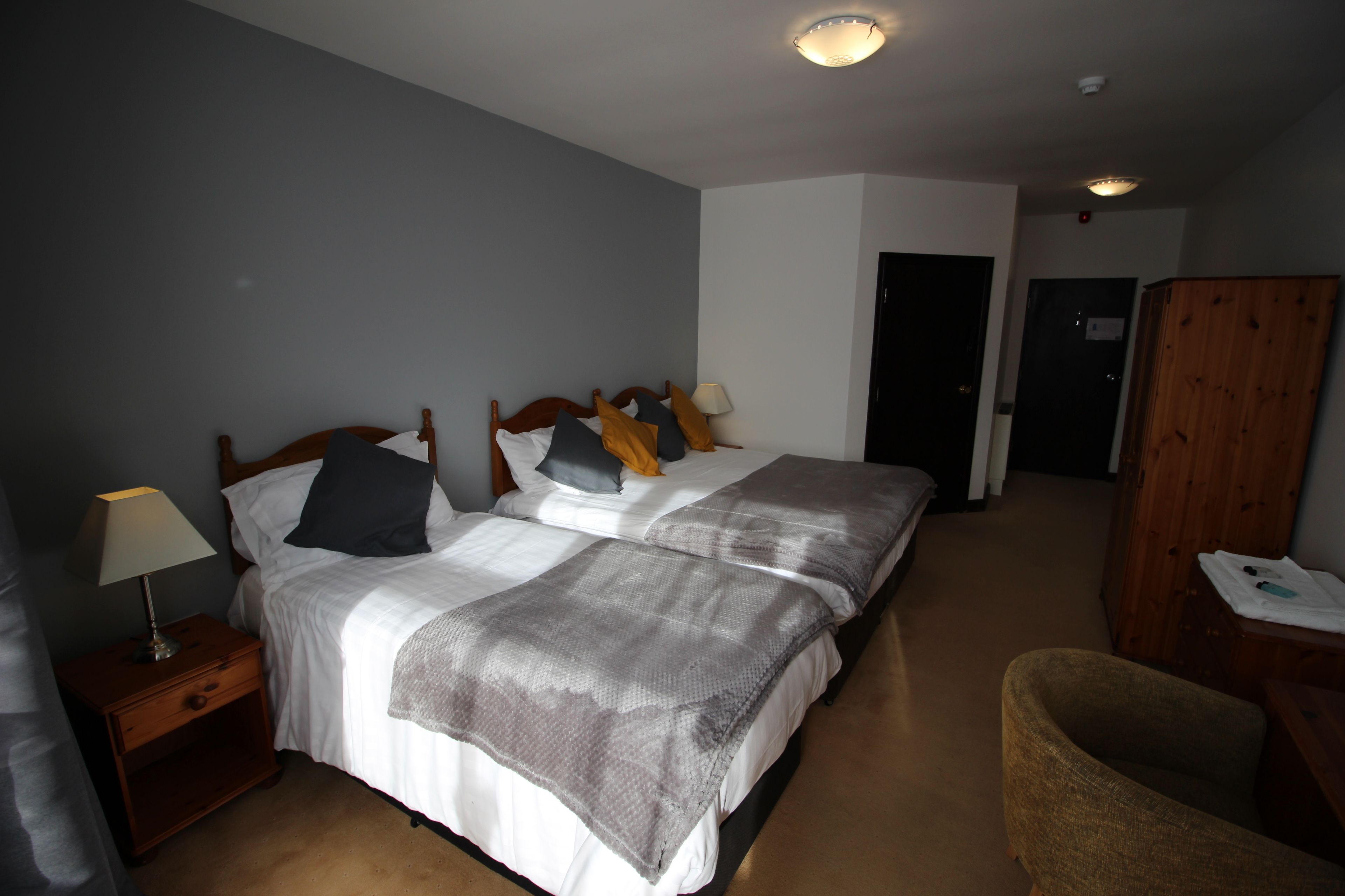 A hotel room with two beds, a nightstand with a lamp, a wooden wardrobe, and a chair.
