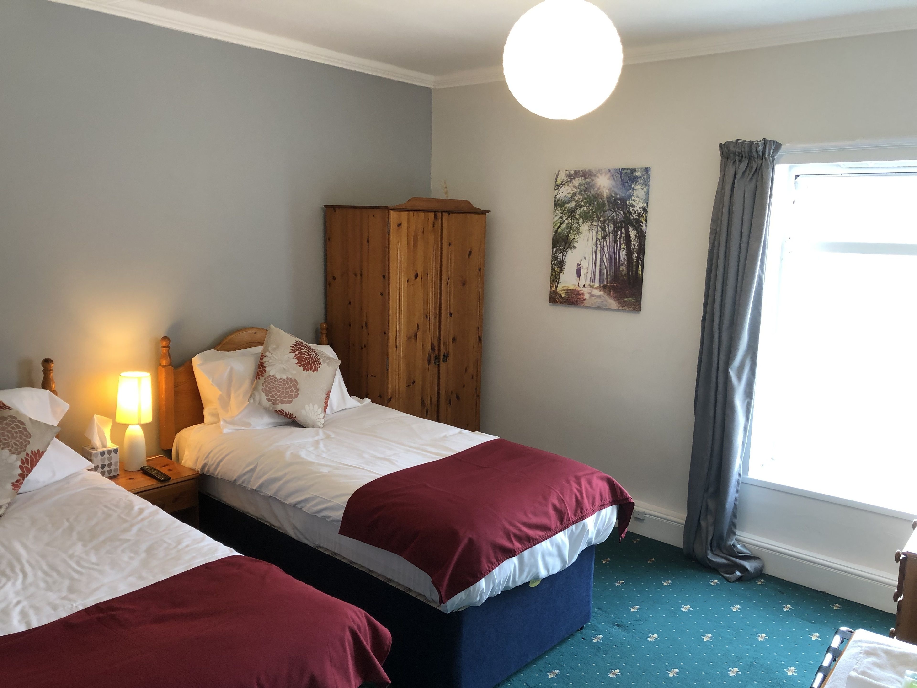 A bedroom with two beds, a wooden wardrobe, a painting, and a window with curtains.