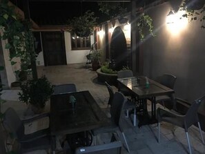 Een terras buiten met een tafel en stoelen, omringd door potplanten en een muur met hangende lampen.