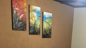 A hotel room with two beds, each with blue bedding and pillows, and a small bedside table. There are three abstract paintings on the wall above the beds. - Hostal Tía Mema (Santa Ana)