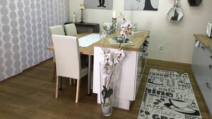 Comedor con mesa de madera, sillas blancas, un jarrón con flores y decoración en la pared.
