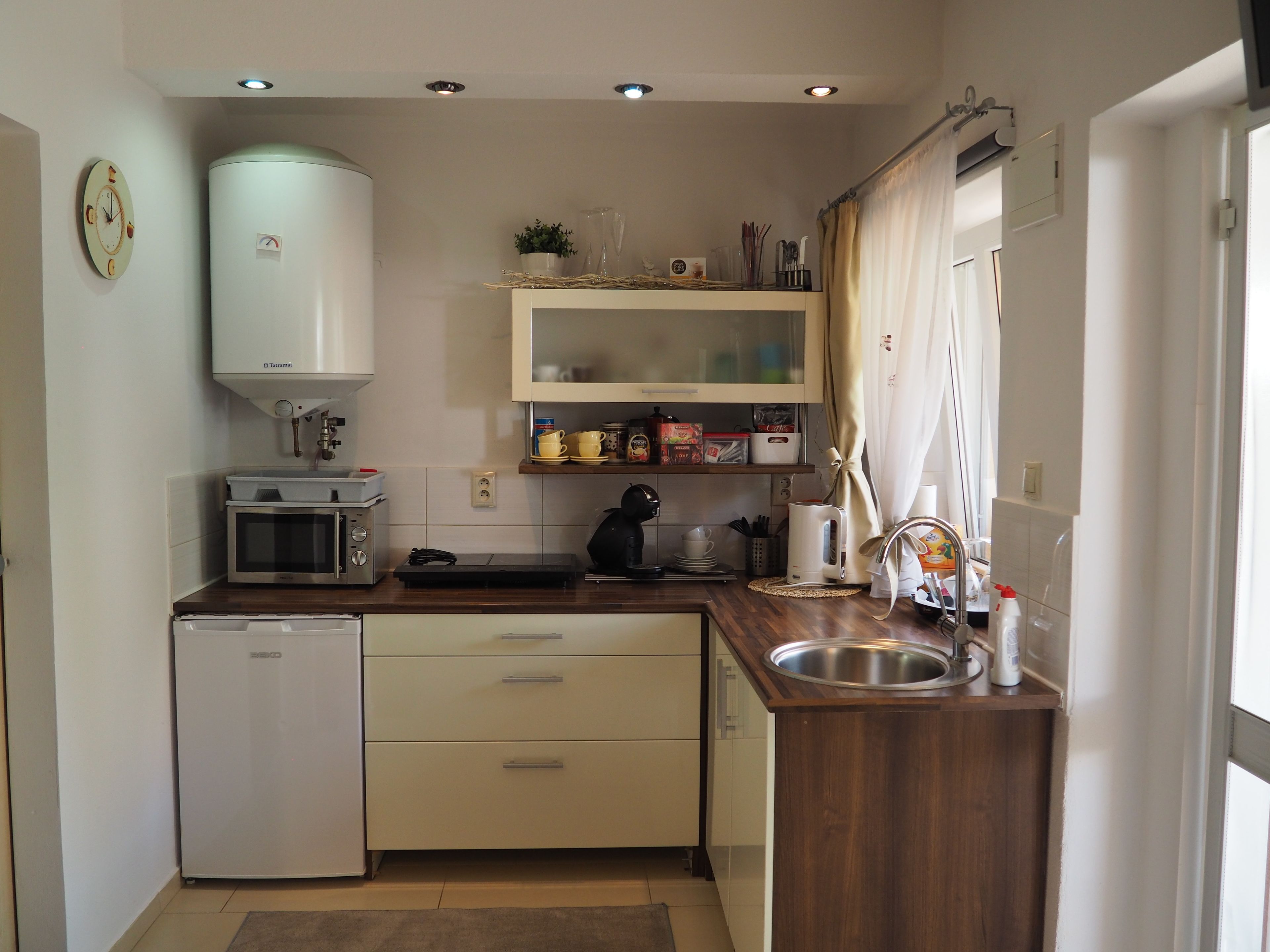 A compact kitchen with a refrigerator, microwave, and sink. There are cabinets above the counter and a small window with curtains.