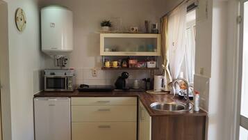 A compact kitchen with a refrigerator, microwave, and sink. There are cabinets above the counter and a small window with curtains.