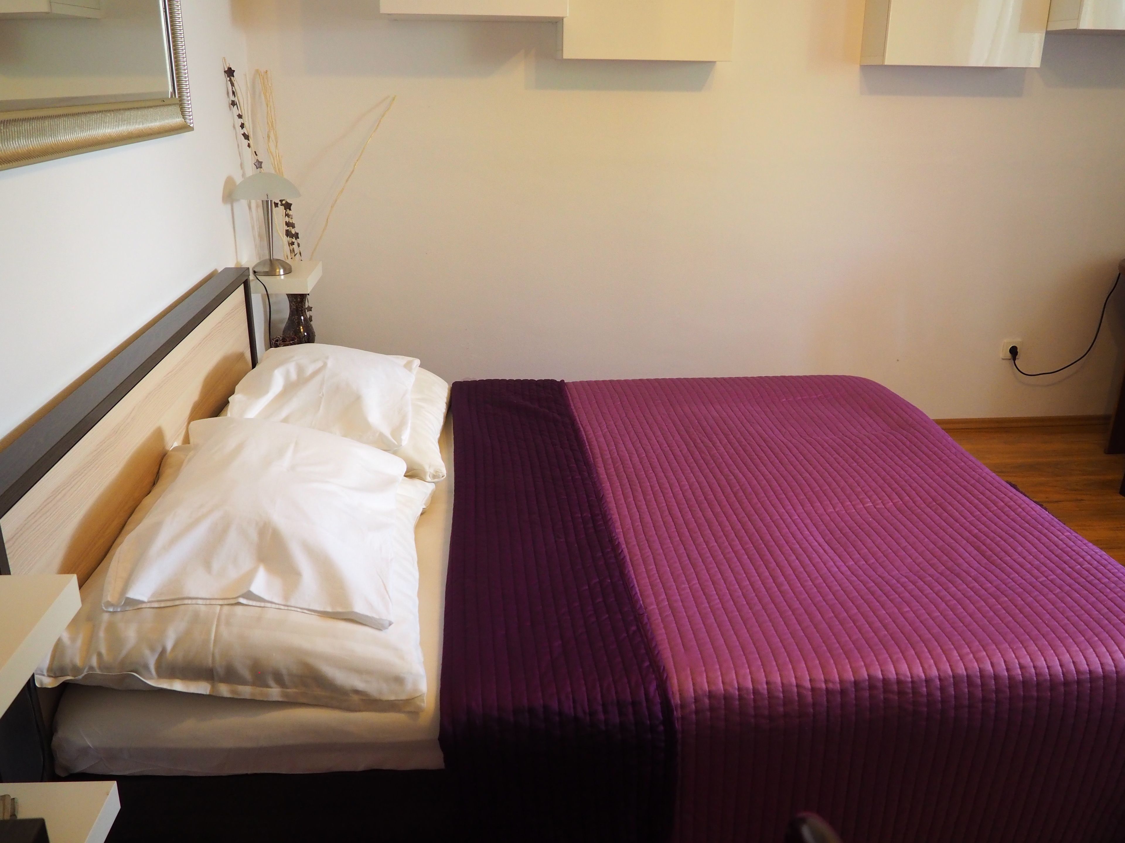 Una cama bien hecha con ropa de cama blanca y morada, una lámpara de noche y un espejo adosado a la pared.