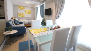 A modern living room with a sofa, a dining table with a yellow and white tablecloth, and a TV mounted on the wall. - KIKU'S ROOM (Osaka)