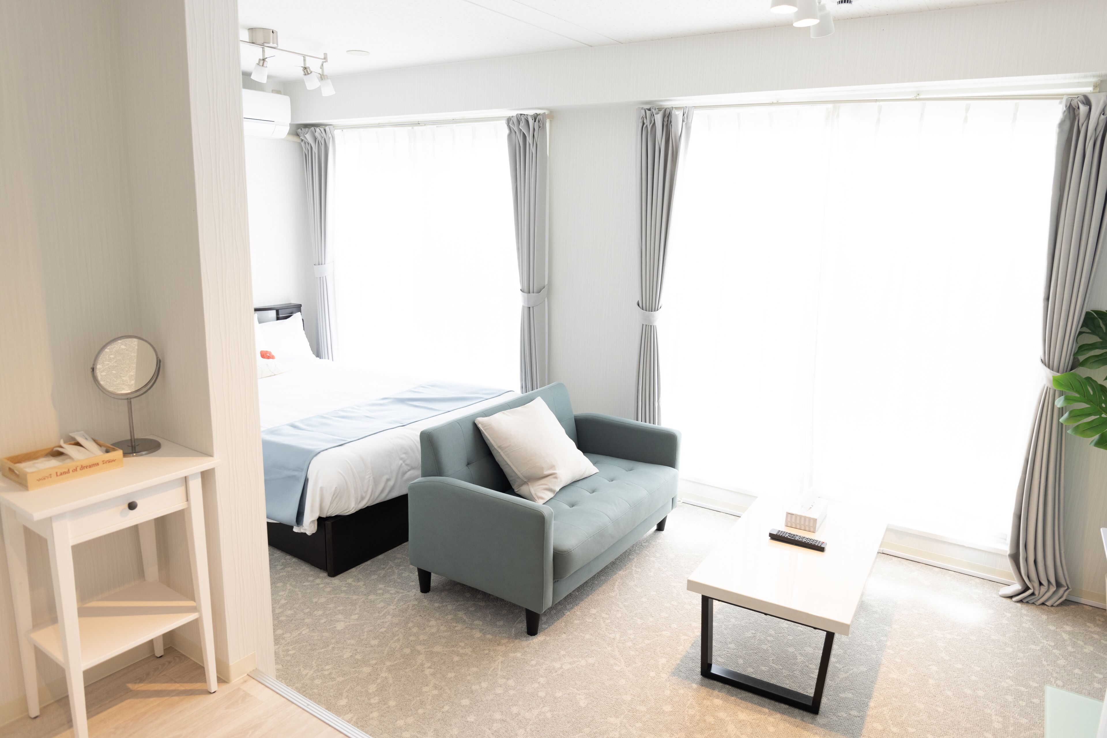 A modern hotel room with a bed, a grey sofa, a small table, and a bedside table with a lamp and a book.