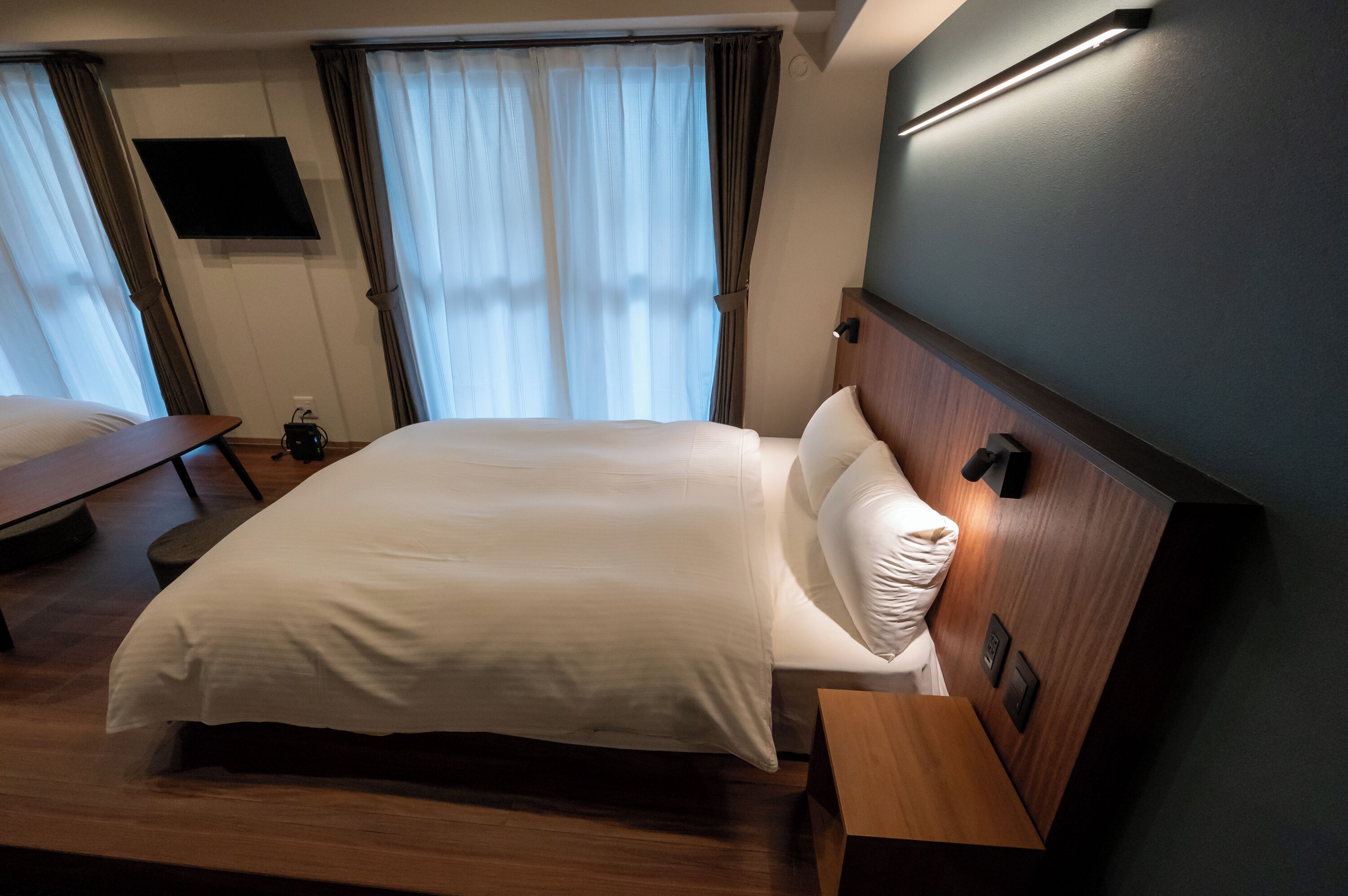 Ein Hotelzimmer mit einem großen Bett, einem Nachttisch, einem flachbildfernseher an der Wand und einem Fenster mit Vorhängen.