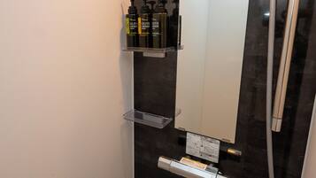 A modern bathroom with a dark tiled wall, a large mirror, a handheld showerhead, and a shelf with toiletries.