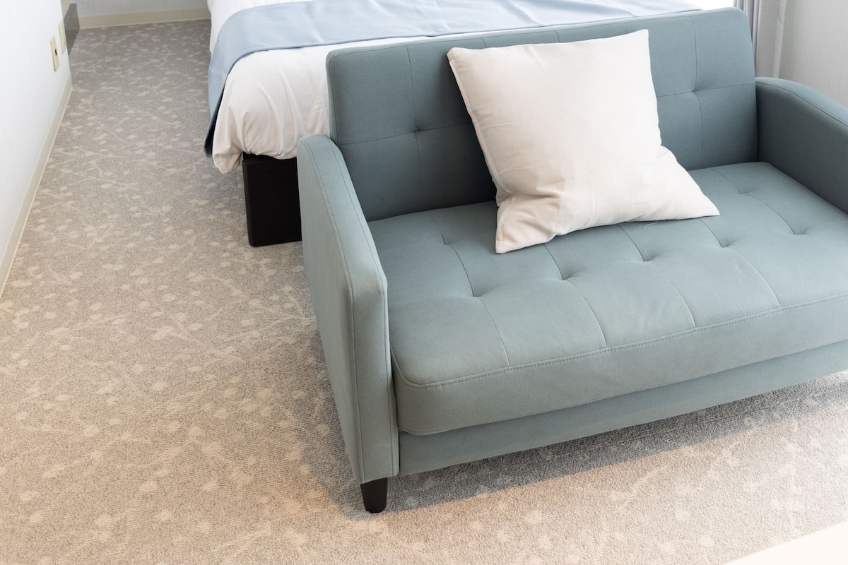 A bedroom with a bed, a blue armchair, a white air conditioning unit, and a window with curtains.