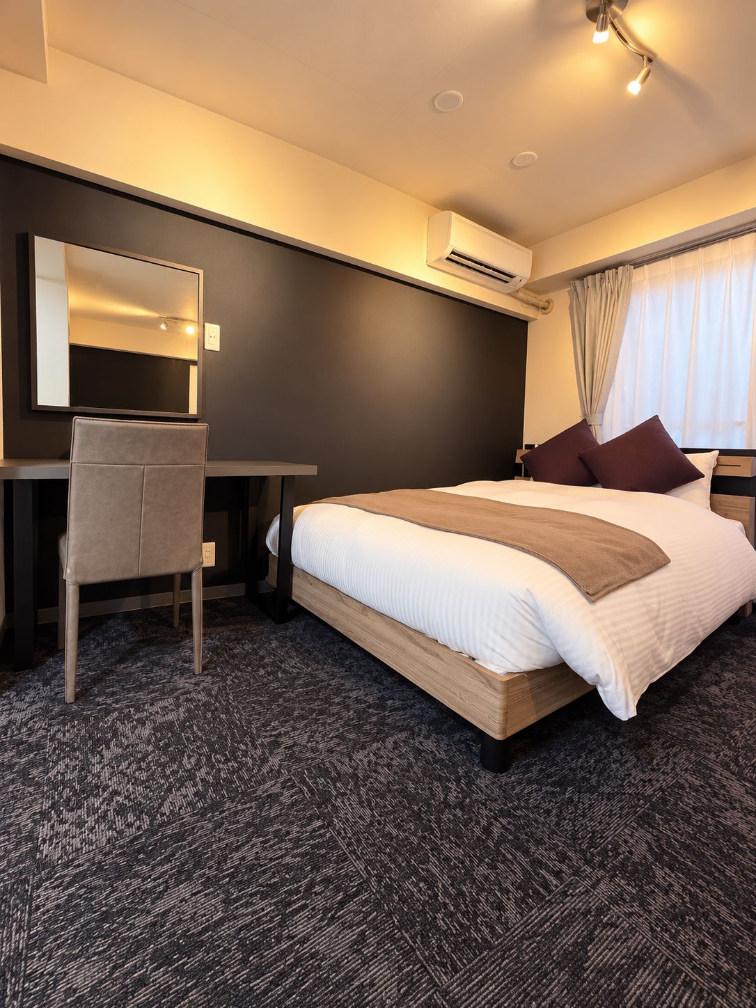 A hotel room with a bed, a desk, a chair, an air conditioning unit, and a window with curtains.