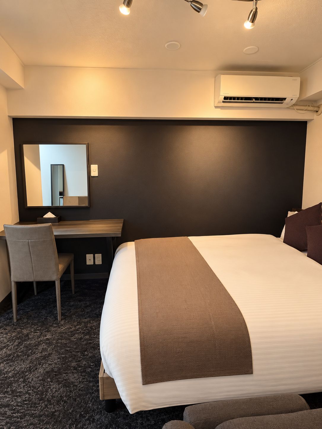 A hotel room with a bed, a desk, a chair, a mirror, and an air conditioning unit.