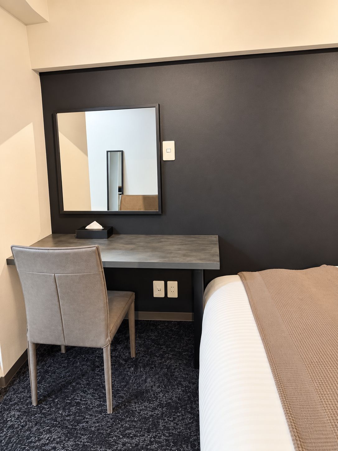 Une chambre d’hôtel comprenant un lit, un bureau avec une chaise, un grand miroir et un interrupteur fixé au mur.