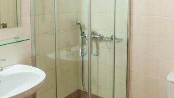A glass-enclosed shower stall with a handheld showerhead, a glass shelf above the sink, and a toilet to the right.