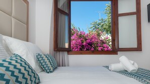 A bedroom with a bed, pillows, a window with a view of pink flowers, and a wooden headboard. - Pallazzo Fortezza (Rethymno)