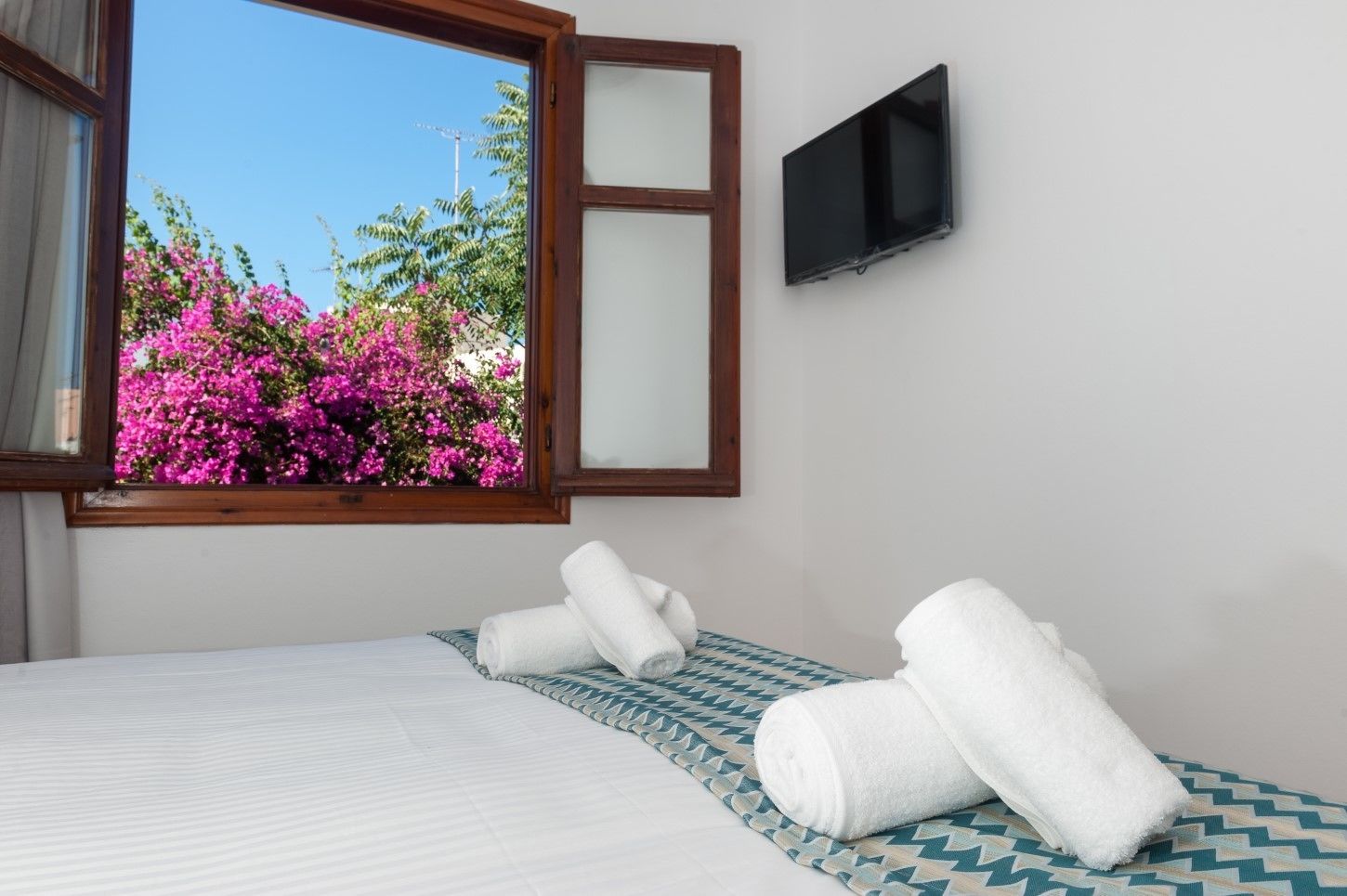 A bedroom with a bed, a window with a view of pink flowers, a flat-screen TV on the wall, and a patterned bedspread.