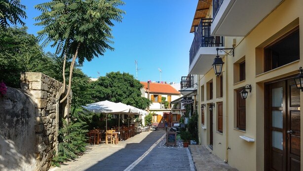 A narrow street with outdoor seating, stone walls, and traditional buildings. - Pallazzo Fortezza (Rethymno)