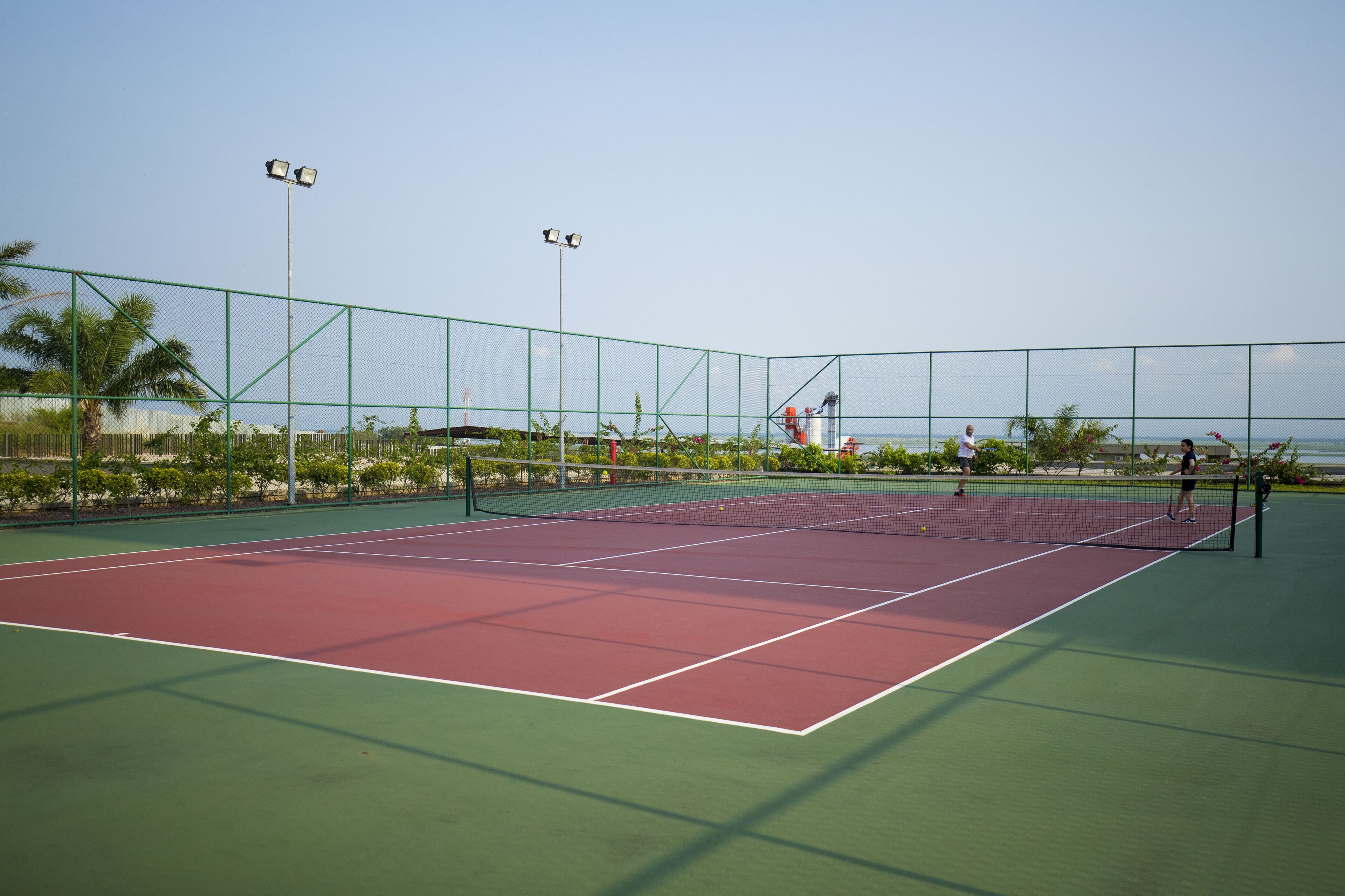 Una cancha de tenis al aire libre con cerca verde, red y cielo despejado.