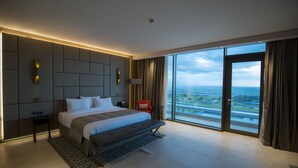 Une chambre d’hôtel moderne avec un grand lit, une banquette, une table de chevet, une lampe et une vue sur la ville grâce à de grandes fenêtres.