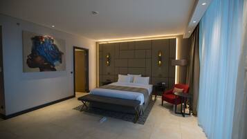 Quarto de hotel moderno com uma cama grande, um banquinho, uma cadeira vermelha e um quadro na parede.
