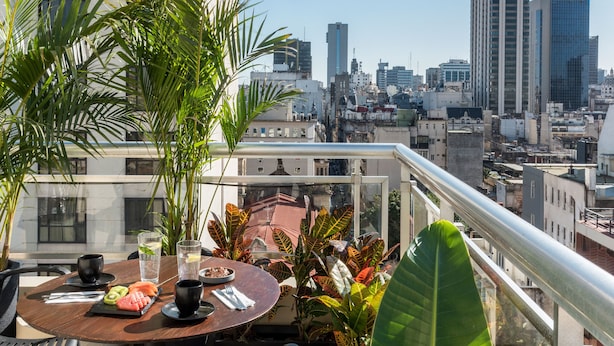 A rooftop terrace with a table set for dining, surrounded by tropical plants, overlooking a city skyline.
