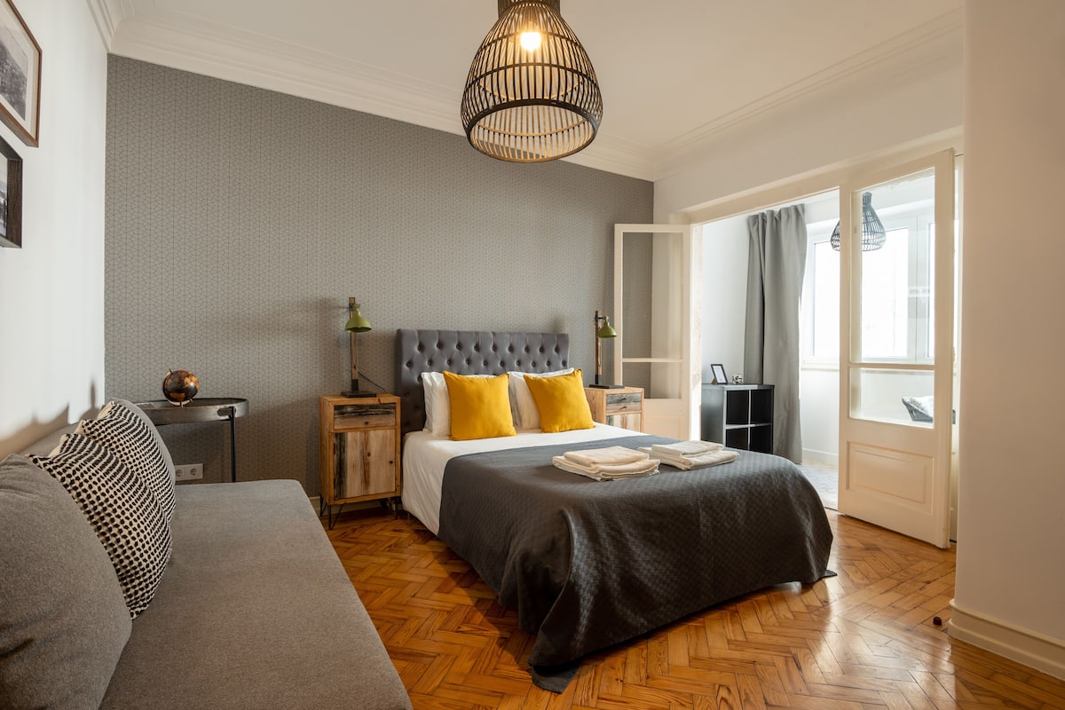 A bedroom with a grey upholstered sofa, a tufted headboard bed with yellow pillows, a wooden floor, and a large window with curtains.