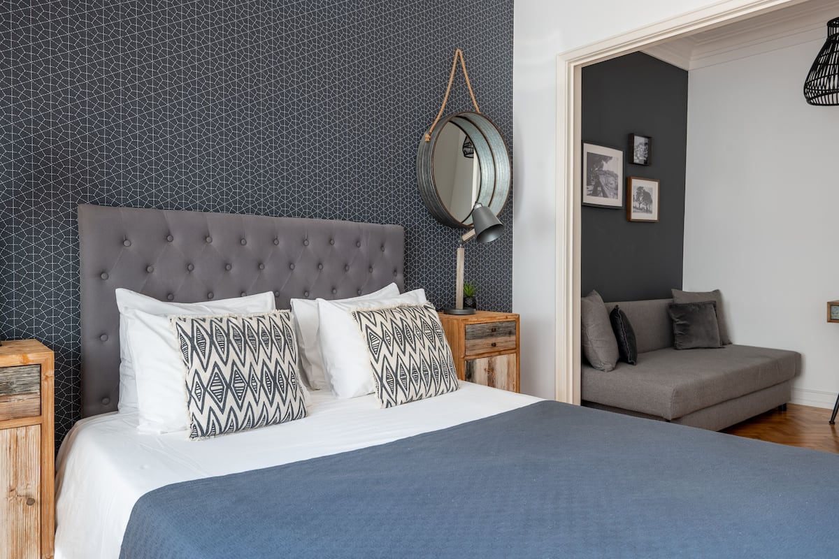 A neatly arranged bedroom with a tufted headboard, a bed with patterned pillows, a bedside table with a lamp, and a grey sofa in the adjacent room.