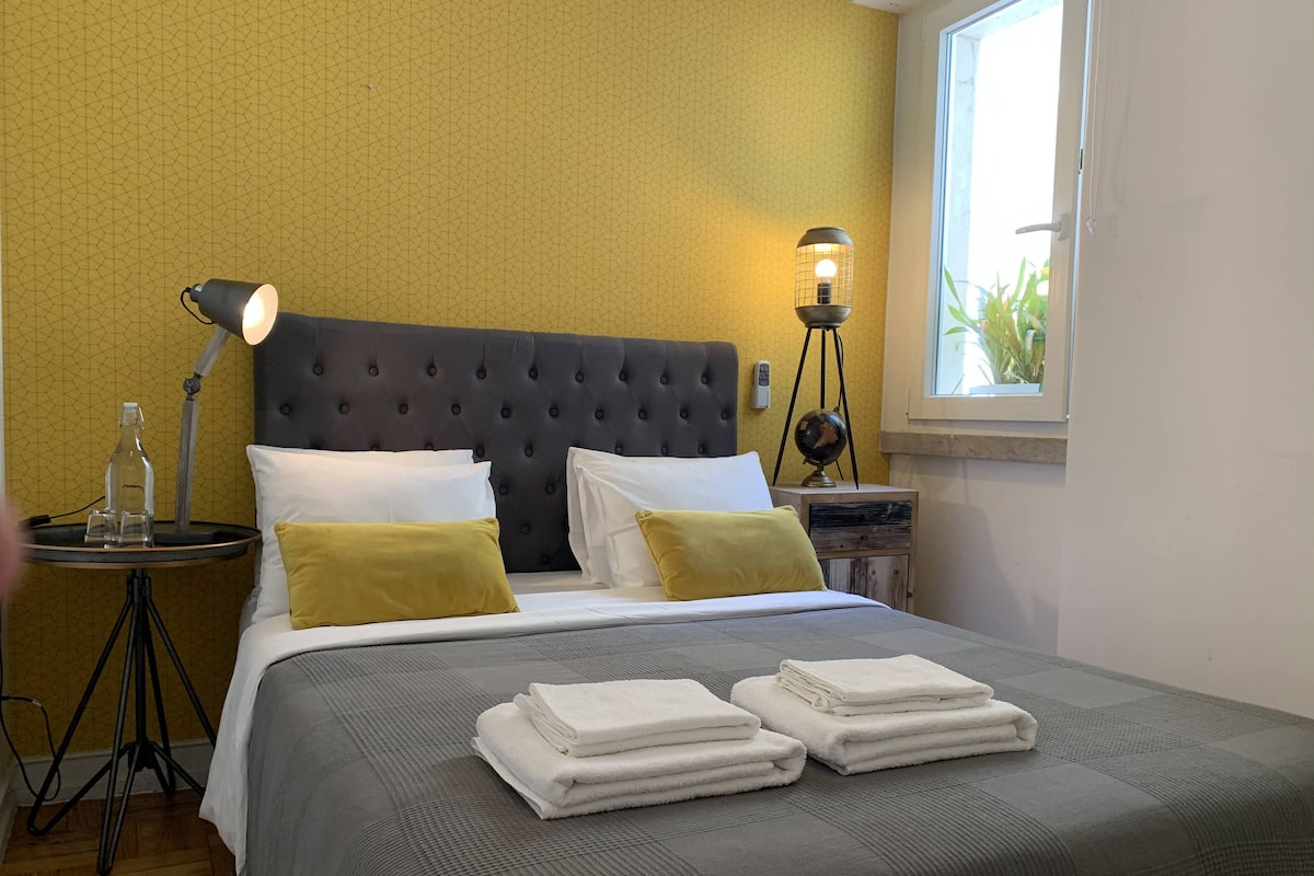 A neatly made bed with white and yellow pillows, a bedside lamp, and a stack of white towels.