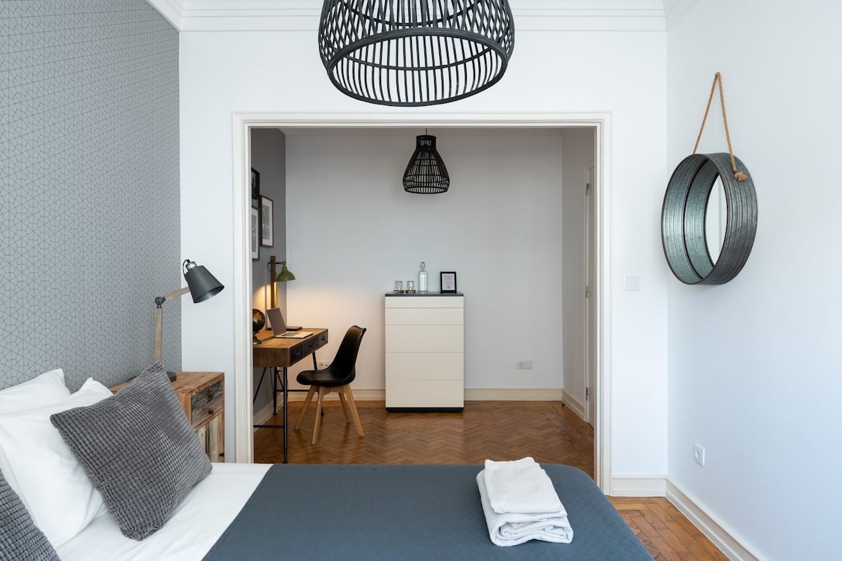 A bedroom with a bed, bedside table, lamp, and a desk with a chair. There is a wardrobe and a hanging light fixture.