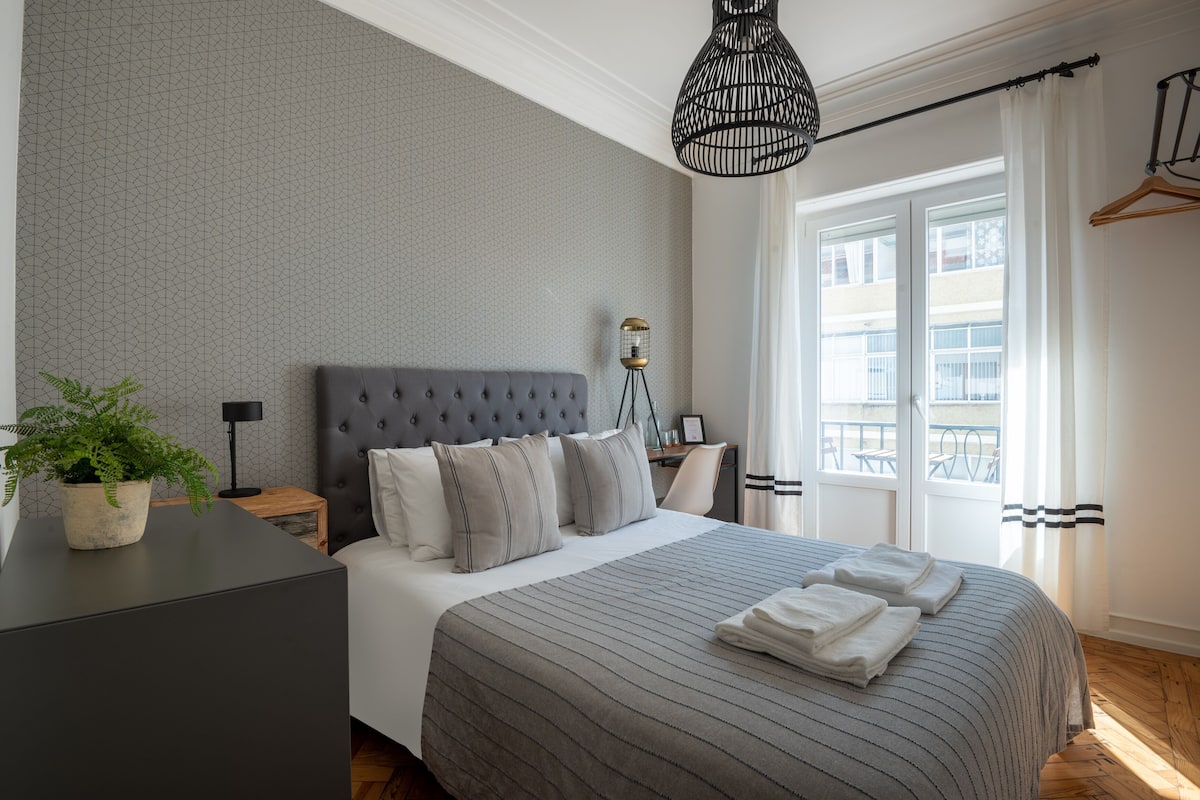 A modern bedroom with a tufted headboard, a large bed with striped bedding, a bedside table with a lamp, a potted plant, and a view of the outside balcony through a window.