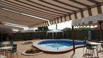 A covered outdoor swimming pool area with lounge chairs and tables, surrounded by a low wall and a pergola with striped awnings.