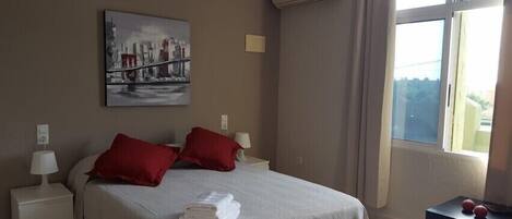 A hotel room with a bed, two red pillows, a white blanket, a folded towel, a nightstand with a lamp, a painting on the wall, and a window with a view.