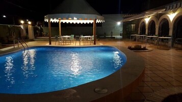 Piscina al aire libre con zona de estar cubierta y mesas, rodeada por un edificio con arcos y suelo de baldosas.