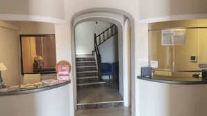 Zona de recepción de hotel con una escalera que sube a otro piso, un mostrador de recepción y un letrero que indica la ubicación del detector de alarma.