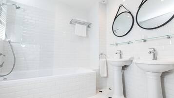 Un baño moderno con ducha de vidrio, una bañera blanca, dos lavamanos pedestal y dos espejos fijos en la pared.
