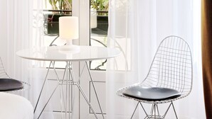 A modern room with a round table and two chairs, a large window with sheer curtains, and a view of greenery outside.
