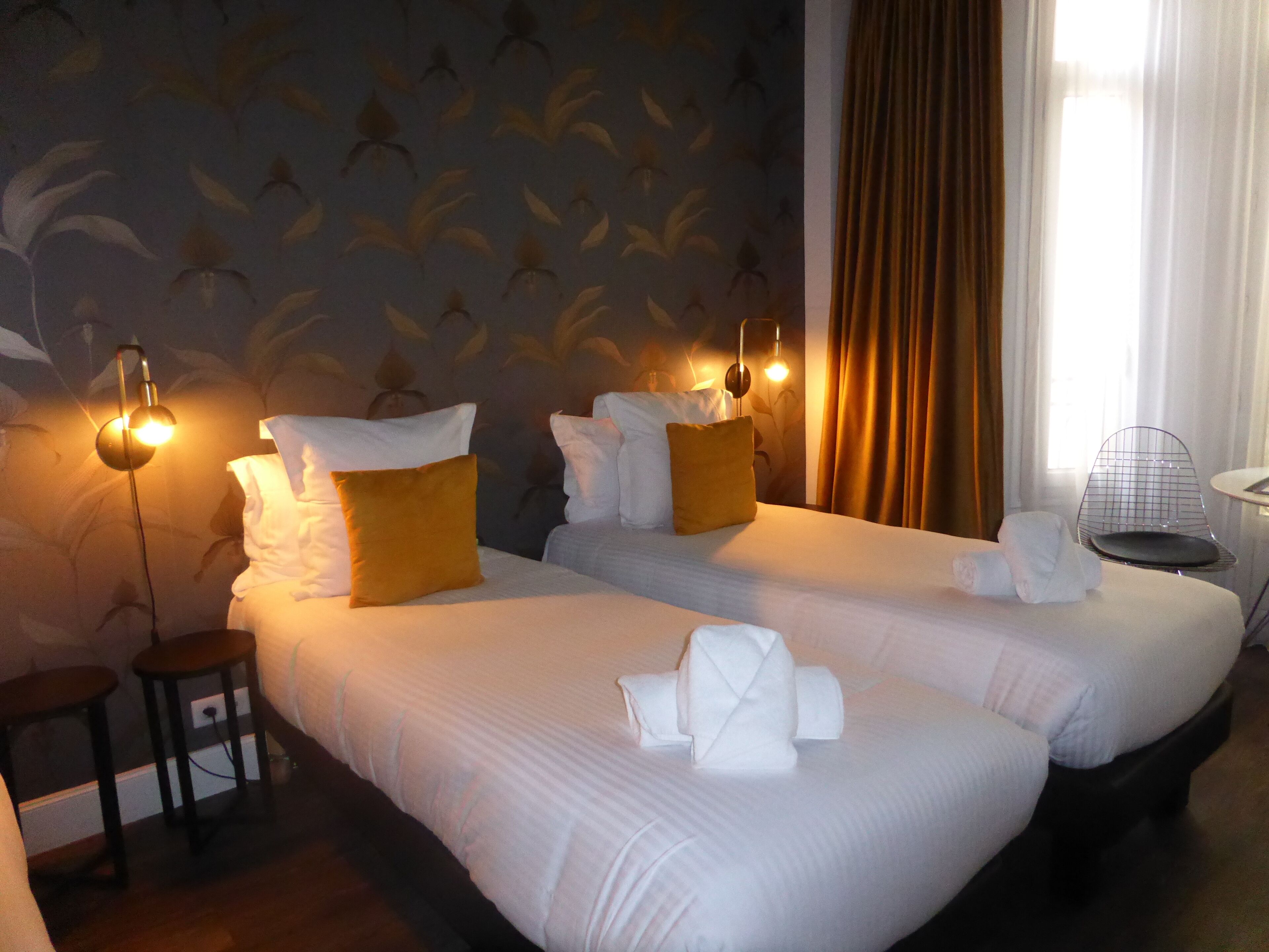 A hotel room with two beds, a small table, a chair, and a wall with a leaf pattern.