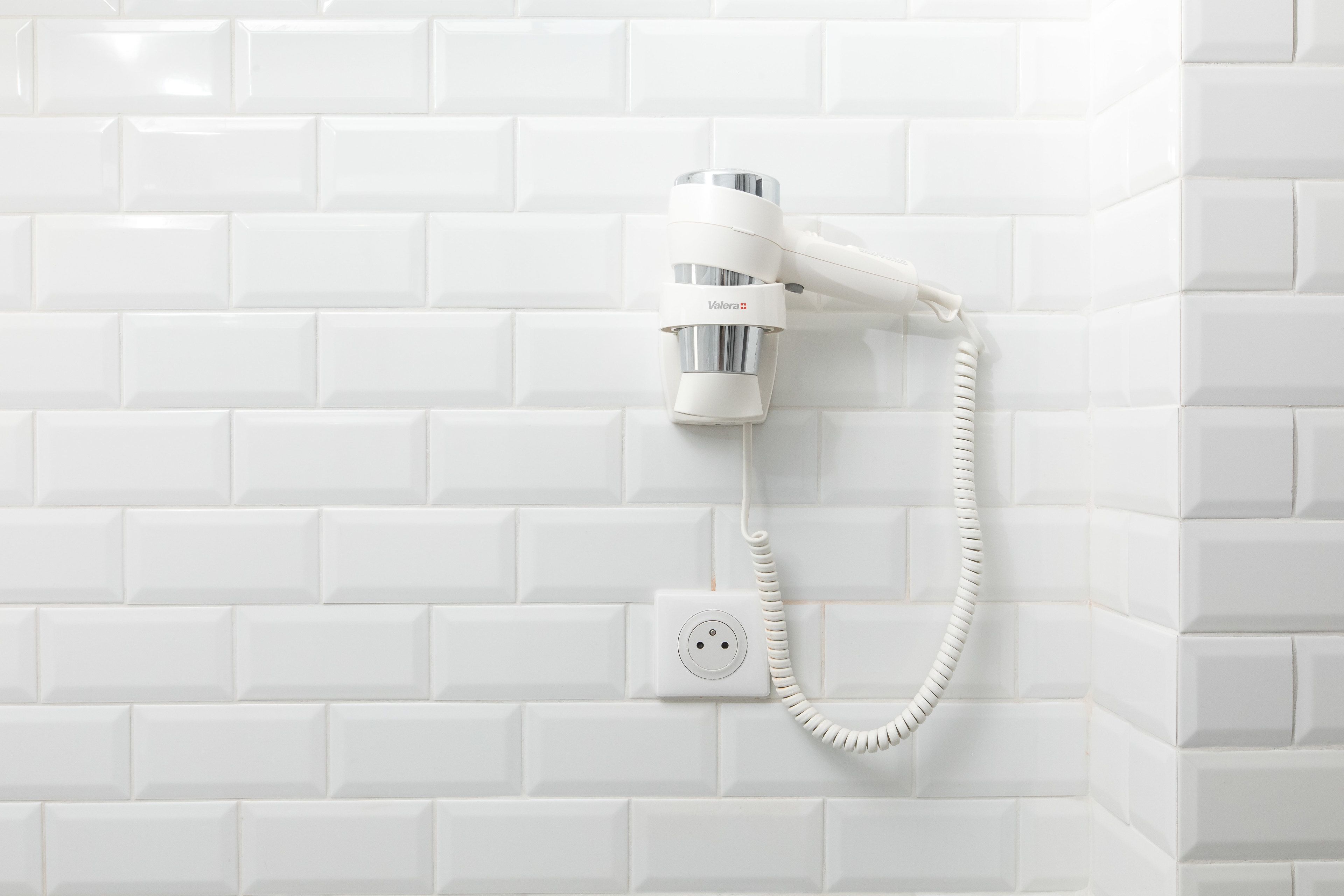 Un sèche-cheveux blanc fixé sur un mur recouvert de carreaux blancs.