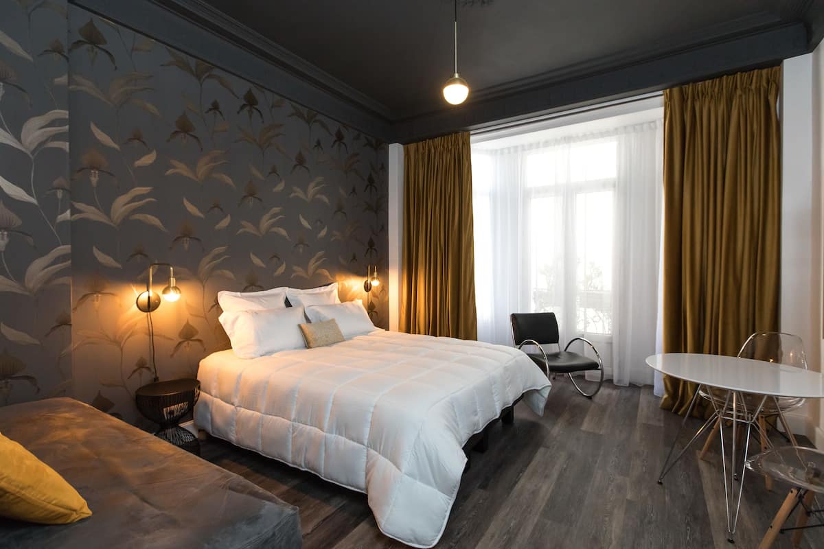 Una habitación de hotel con una cama grande, una mesita de noche, una silla, una pequeña mesa y una ventana con cortinas.