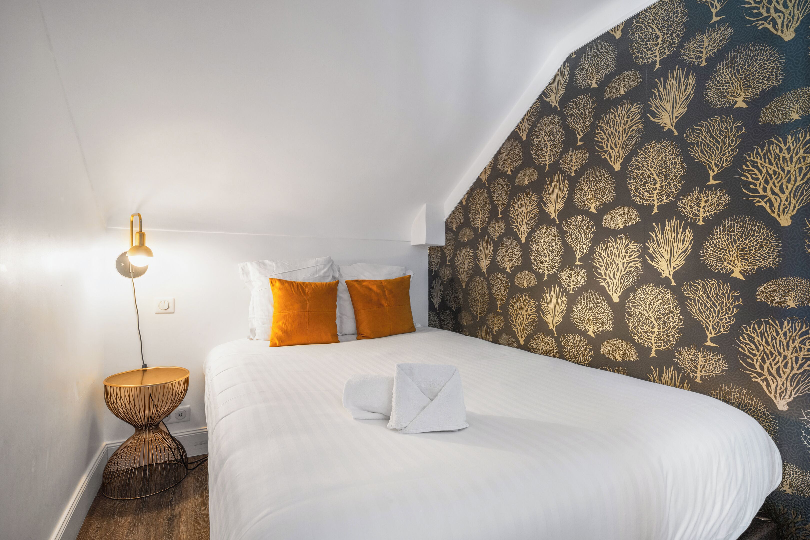 A neatly made bed with white linens and two orange pillows, a bedside lamp, and a patterned wallpaper.