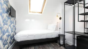 A compact bedroom with a bed, a small stool, a desk, and a shelving unit.
