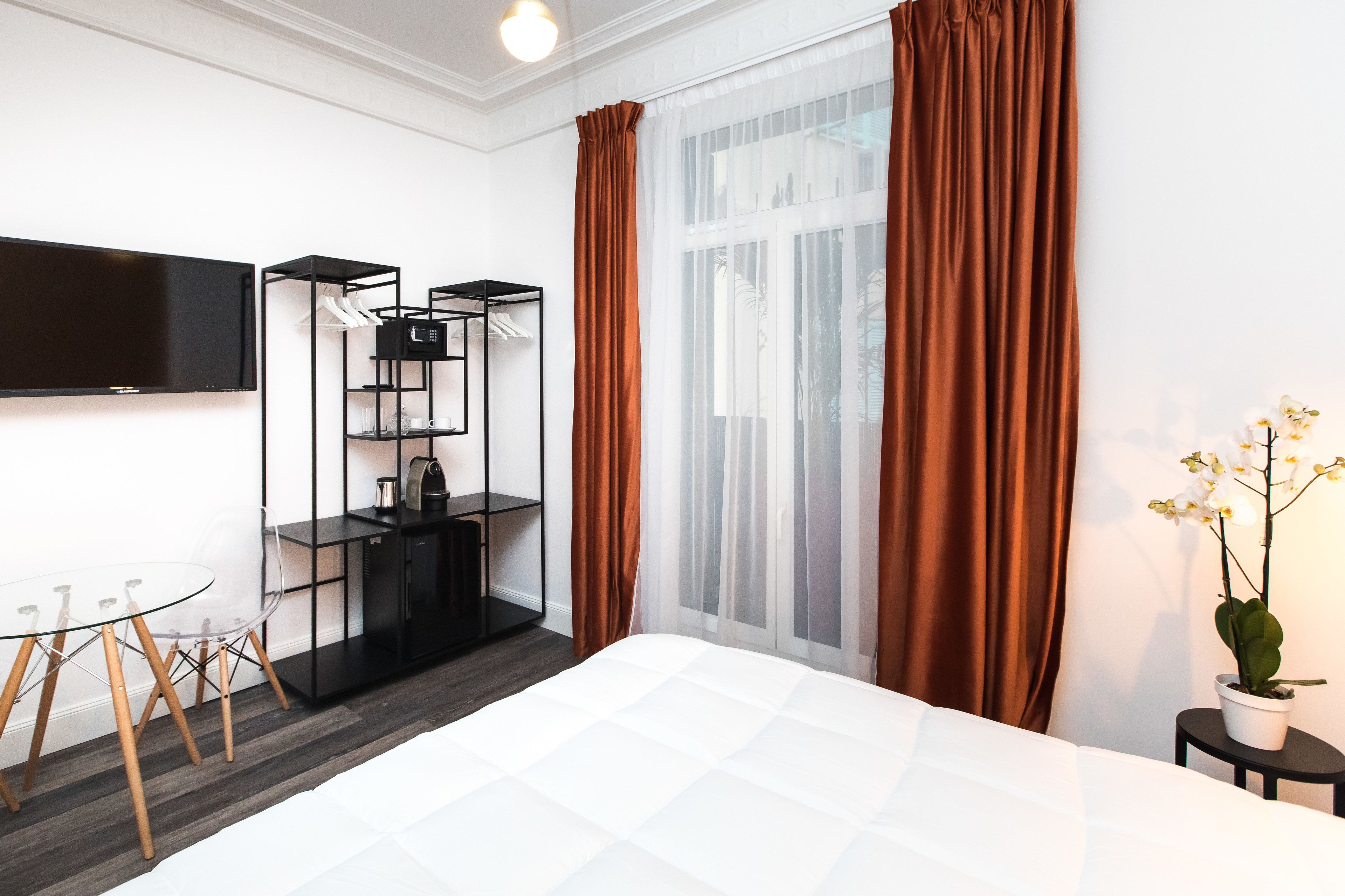 A modern hotel room with a large bed, a TV mounted on the wall, a black shelving unit, and a potted orchid.